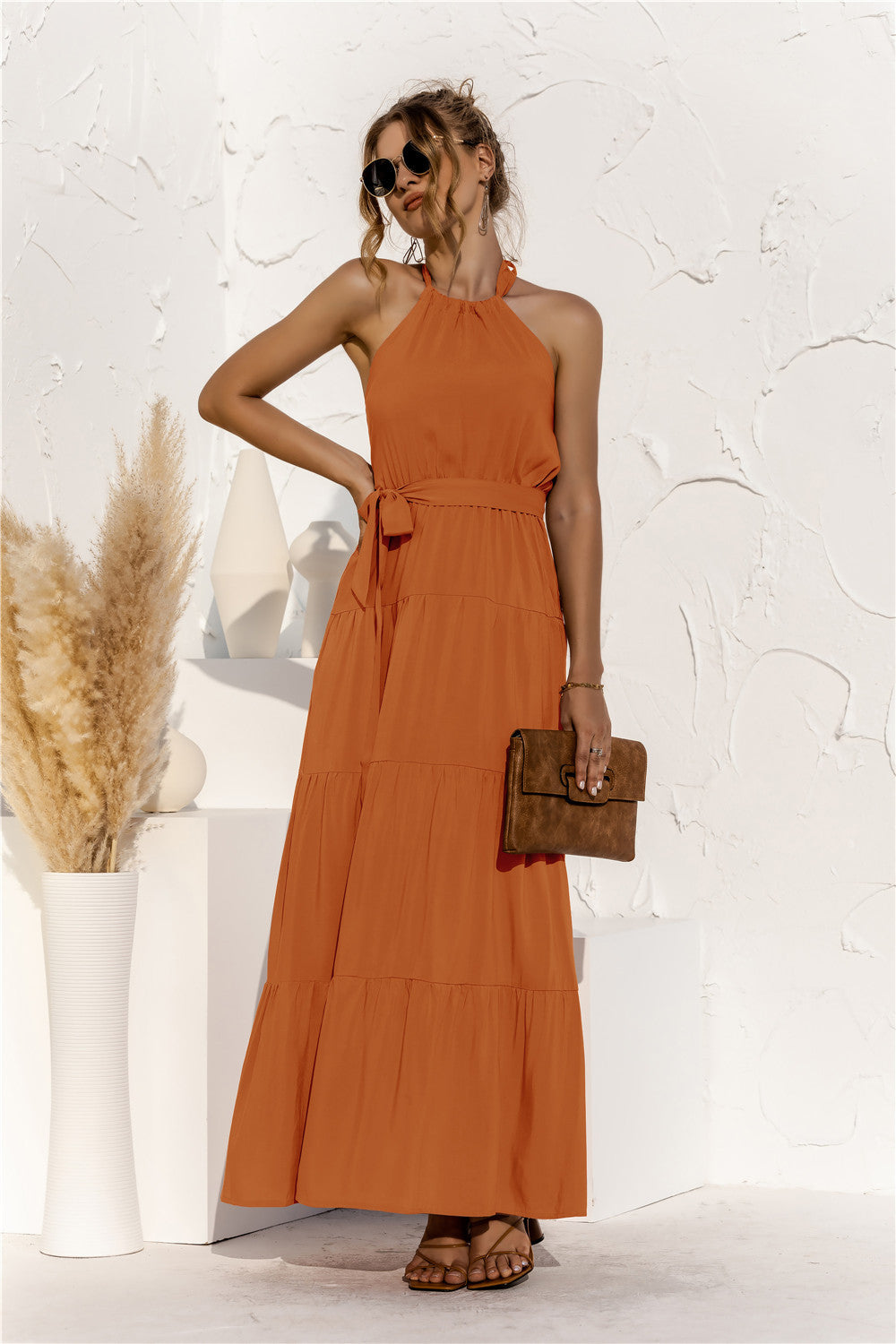 Model is wearing halter neck tiered maxi dress with tie front waist in the color terracotta.
